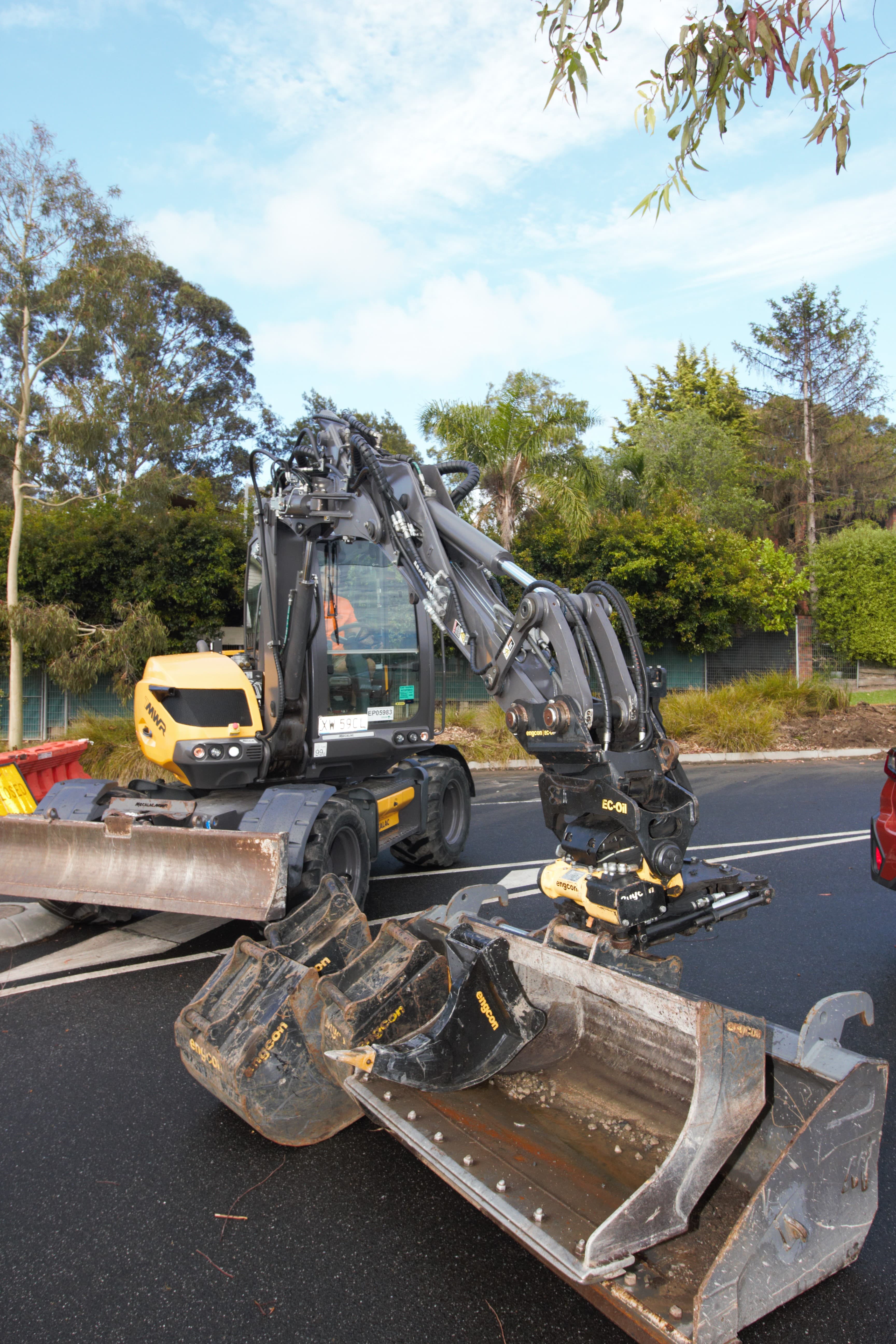 Yellow and black Mecalac MWR wheeled excavator with buckets on asphalt, under cloudy sky.