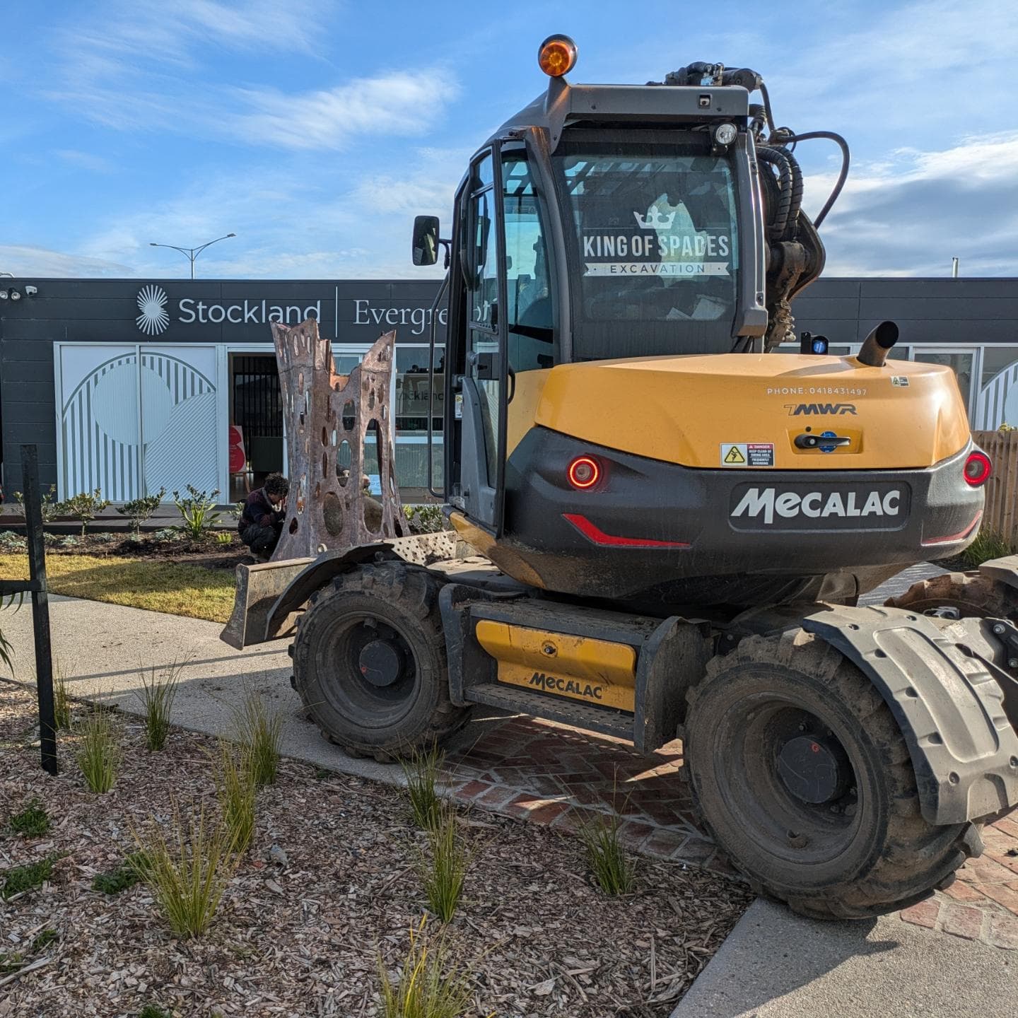 Yellow Mecalac excavator with 'King of Spades' logo, parked by Stockland Evergreen building.