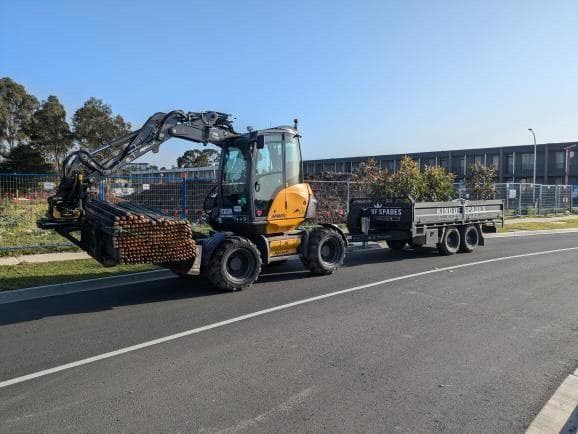 Yellow wheeled excavator carrying brown poles, towing a black trailer on a paved road.